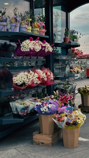 A display of fresh flowers organized on dark shelving at a florist shop. The top shelf features small, colorful potted plants wrapped with pastel ribbons, alongside small bouquets of mixed flowers. Th