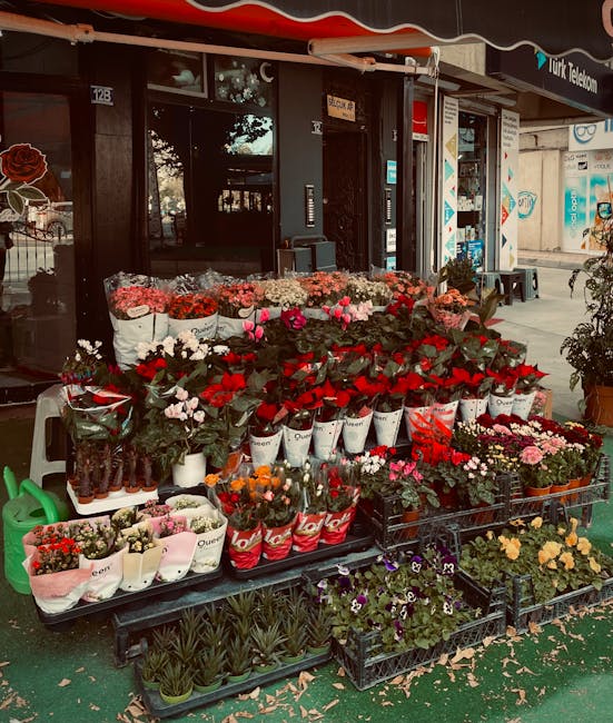 An outdoor display of fresh flowers arranged on black plastic tables in front of a shop on a busy street, showcasing a variety of colorful blooms including red, pink, white, and orange flowers such as