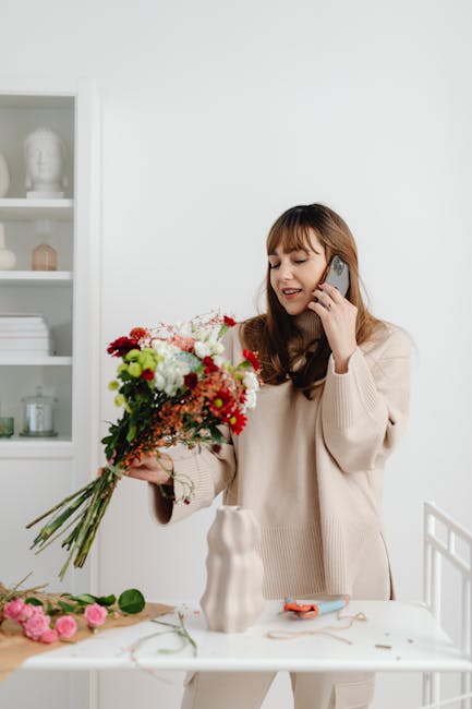 A young woman with light brown hair and bangs, dressed in a beige sweater, is holding a large, vibrant bouquet of fresh flowers including red, white, pink, and green blooms, with visible petals and st