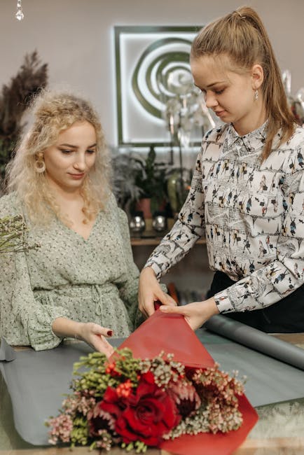 Two women are engaged in floral arrangement in an indoor setting, with a bouquet of vibrant red roses, pink and white carnations, and greenery wrapped in red paper placed on the table before them. The