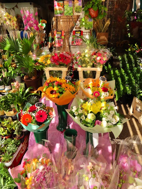 A vibrant display of fresh flower bouquets arranged on a pink table in a floristry shop. The bouquets include bright pink, orange, yellow, and white flowers, such as gerberas and roses, wrapped in col