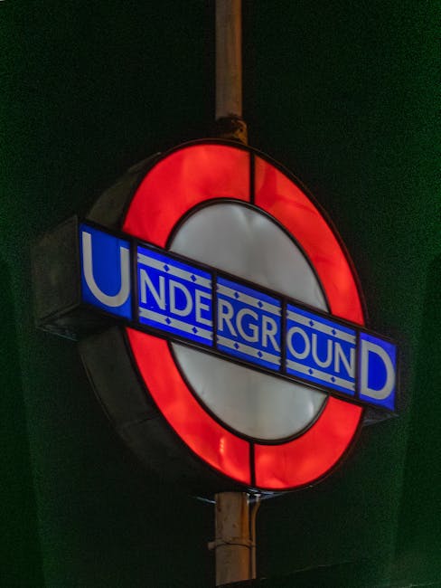 A circular underground railway sign illuminated at night, featuring a red outer ring, a white central background, and a blue horizontal bar with the word 'UNDERGROUND' in white block letters. The sign