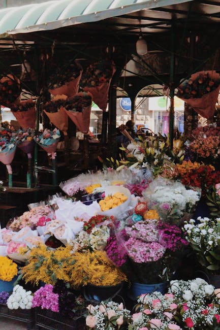 An outdoor flower market stall under a curved, green canopy displaying a vibrant assortment of fresh floral arrangements. The stand features numerous bouquets and bunches wrapped in brown craft paper 
