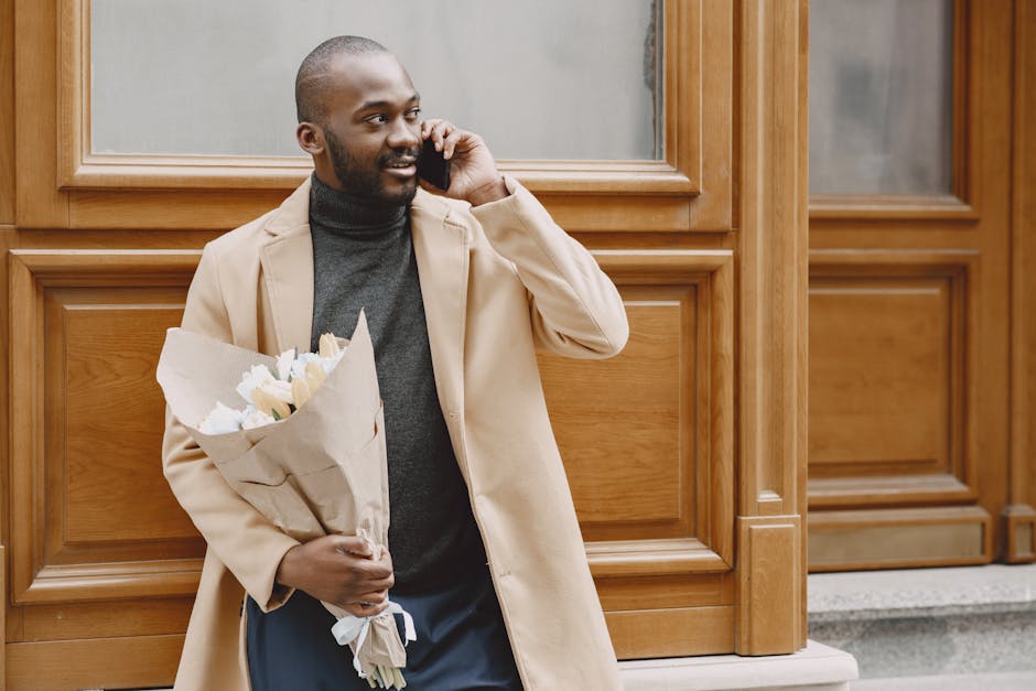 A man with dark skin, a neatly trimmed beard, and a buzz haircut is standing outside in front of a wooden-paneled building. He is wearing a beige overcoat over a dark gray turtleneck sweater. He holds