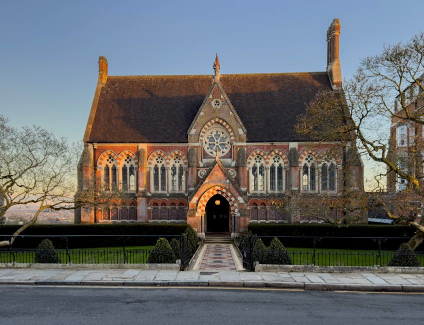 A large, historic stone church building with Gothic architectural features, including pointed arches and a circular stained glass window above the entrance, set against a clear blue sky during late af